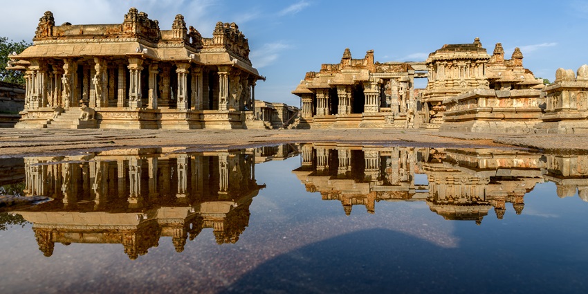 Stone temple ruins with tall pillars reflected in water at temples in Karnataka.