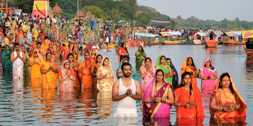 People half dipped in water standing and praying with a few boats parked behind them.