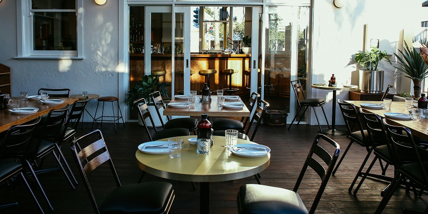 Sunlit dining room with black chairs, round tables, and a bar visible through glass doors
