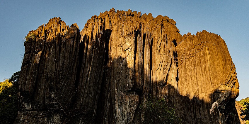 A tall, jagged rock formation with vertical striations is partially illuminated by sunlight against a clear blue sky.
