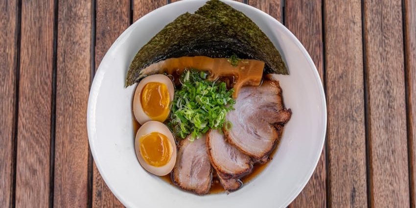 A round bowl of ramen with pork slices, two soft-boiled eggs, green onions, and a sheet of nori.