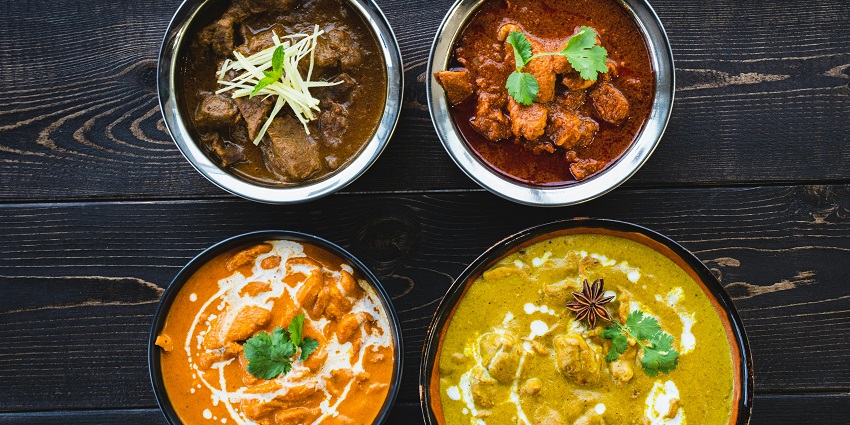 A flat-lay of four different curries in round metal bowls on a dark wooden table.
