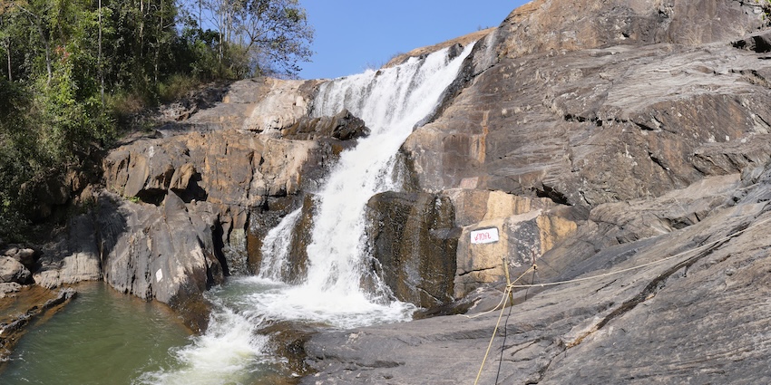 A wide waterfall flows over the edge of a cliff near a wooden viewing deck in a thick forest.