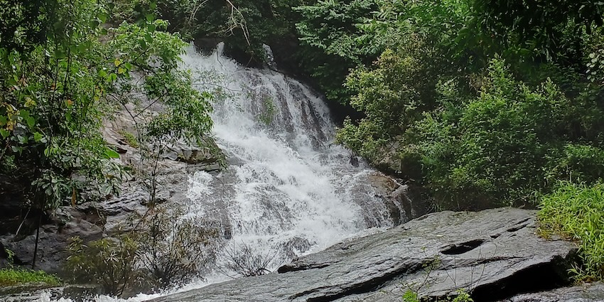 Wide waterfall over layered brown rocks, flowing into a pool, one of the best among Tripura waterfalls.