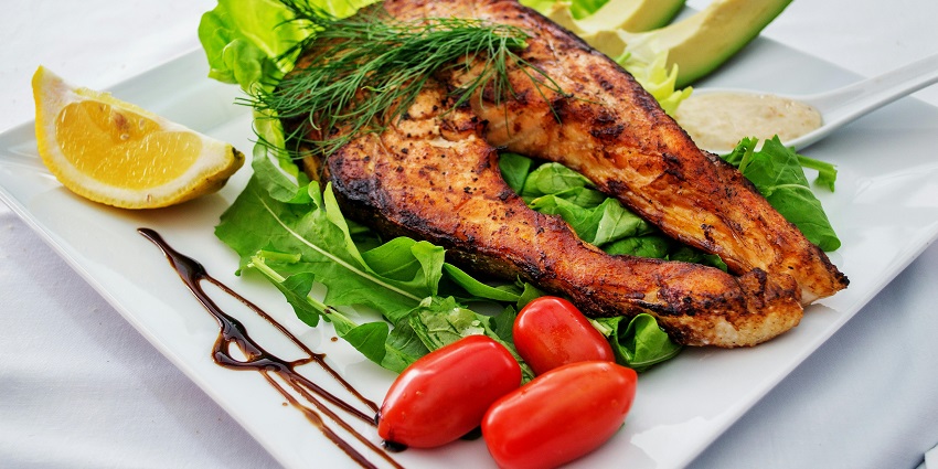 Grilled salmon steak served with fresh greens, avocado slices, lemon, and cherry tomatoes.