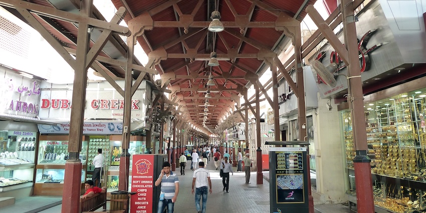 A narrow, crowded alley in the Dubai Gold Souk, lined with brightly lit storefronts.