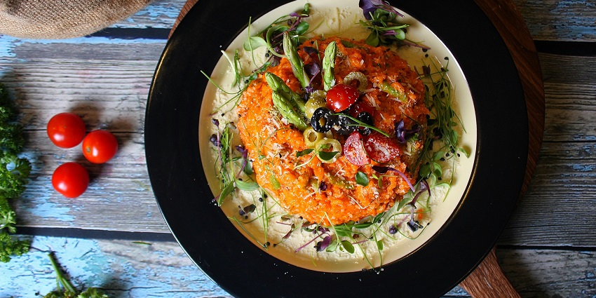 Colourful risotto topped with asparagus, cherry tomatoes, olives, and herbs on a black plate.