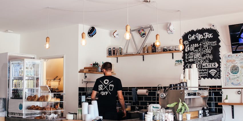 Barista working behind a modern café counter with a chalkboard menu and hanging lights.