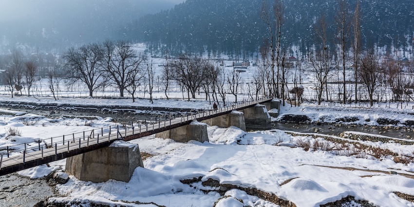 Cars and people passing over a water body on the bridge and surrounding area, showing snowfall in Srinagar.