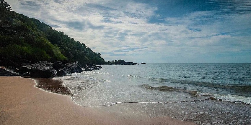 Gentle waves wash onto a sandy beach with a rocky, tree-covered hillside nearby.