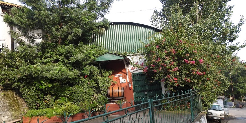 Entrance of Kasauli Brewery surrounded by greenery and flowering plants along a hilly road.