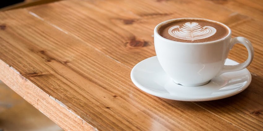 White ceramic cup of cappuccino with leaf latte art placed on a wooden table.