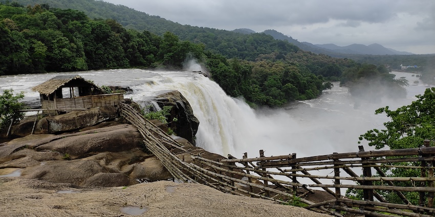Waterfall tumbling over wide, layered brown rock formations under a bright blue sky.