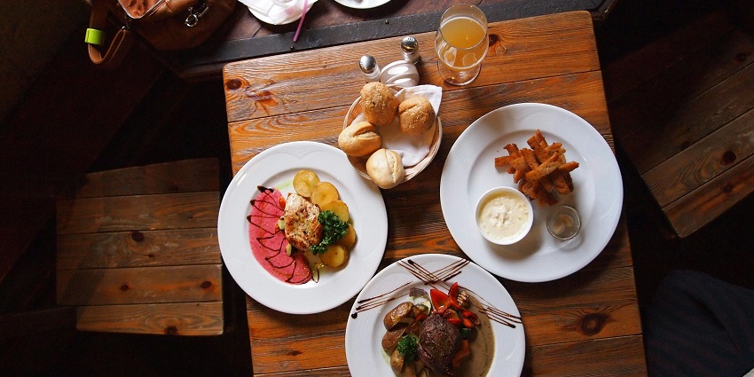 Wooden table set with bread rolls, meat dishes, fries, and a glass of juice for dining.
