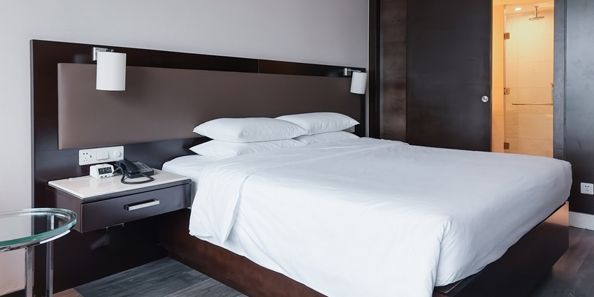 Modern hotel room with clean white bedding, dark wood furniture, bedside table, and sliding door to bathroom