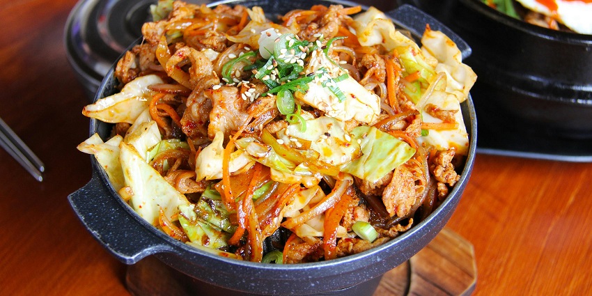 Hot bowl of stir-fried pork with cabbage, carrots, and sesame, garnished with scallions.