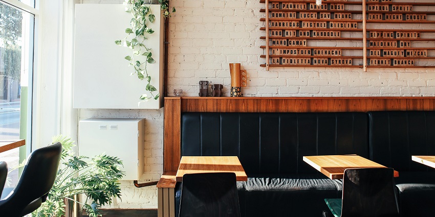 Bright café corner with wooden tables, black seating, indoor plants, and a wall menu board.