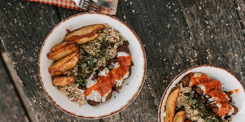 Bowls of rice with falafel, potato wedges, herbs, and sauces on a rustic wooden table.
