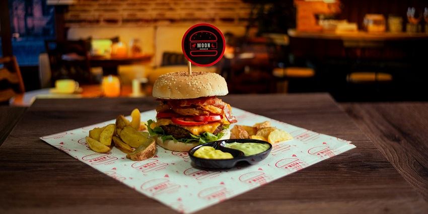 Sesame burger with bacon, tomato, fries, and dips served on a wooden table, one of the popular restaurants in Jorhat.
