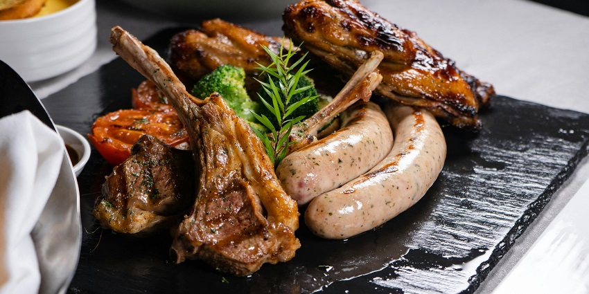 Grilled lamb chops, sausages, and roasted vegetables served on a black stone platter.