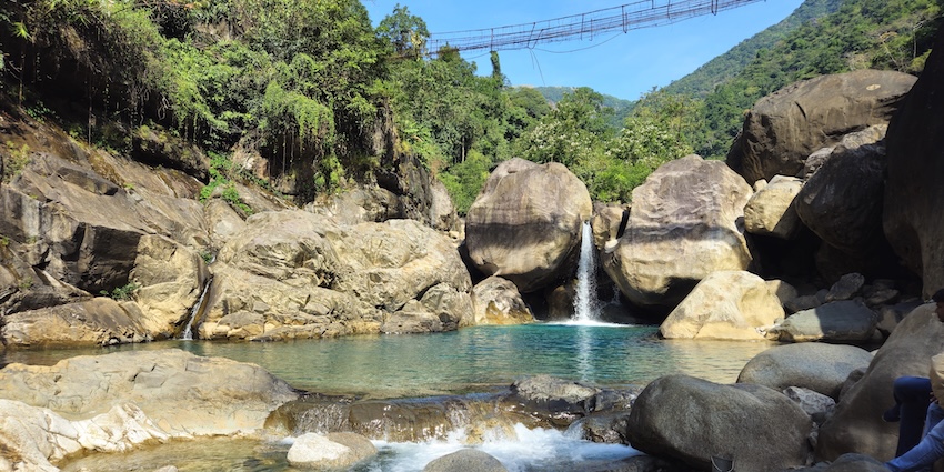 A secluded natural pool with turquoise water, boulders, and a small waterfall in a forest.