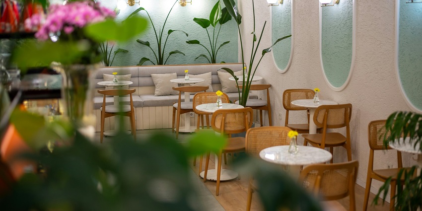 Cosy café with green accents, wooden chairs, marble tables, and fresh plants inside.
