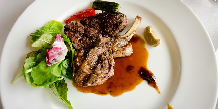 Grilled lamb chops with sauce, greens, and roasted vegetables served on a white plate.
