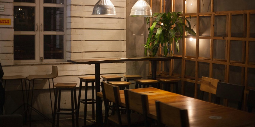 Warmly lit restaurant corner with wooden tables, high stools, hanging lamps, and indoor plants, one of the cosy restaurants in Shillong.
