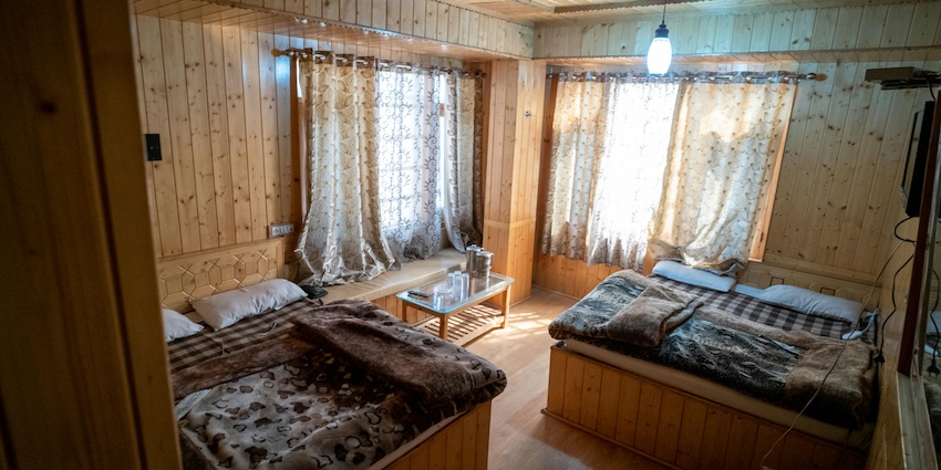 Small wooden beds inside a small room with walls of wooden planks and two windows