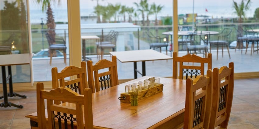 A large dining room with wooden tables and chairs, with a view of an outdoor patio and the ocean.